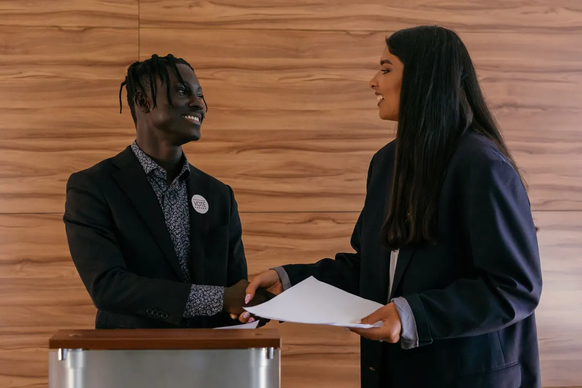 A candidate wearing a vote button shakes hands with a staffer at a lectern