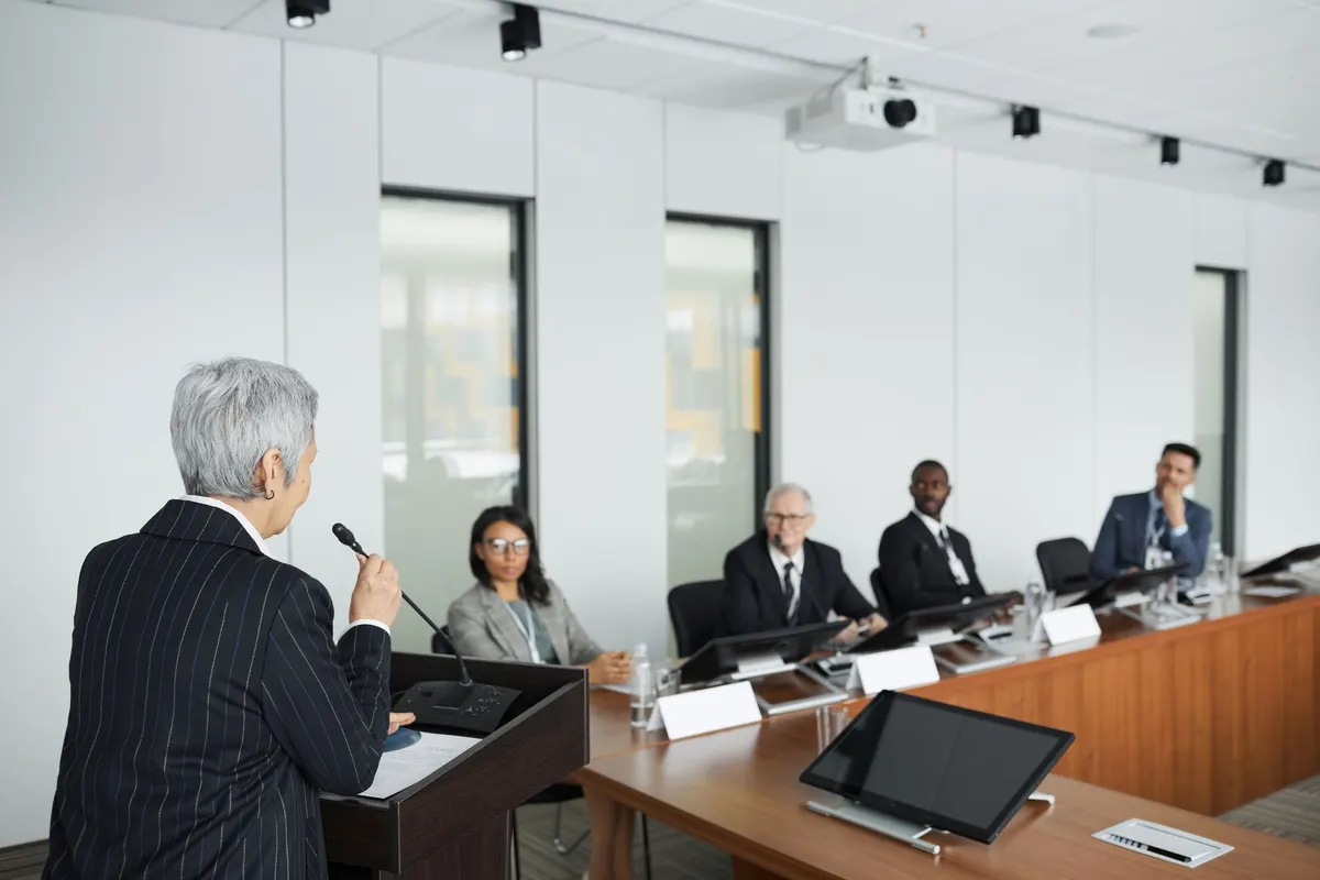 A woman testifies at a lectern before a panel seated at a long committee desk