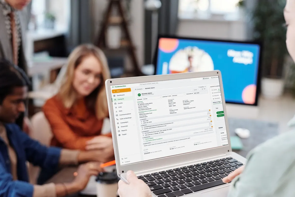 A staffer holds a laptop showing a FundraiserMax contact record while colleagues work at the table behind